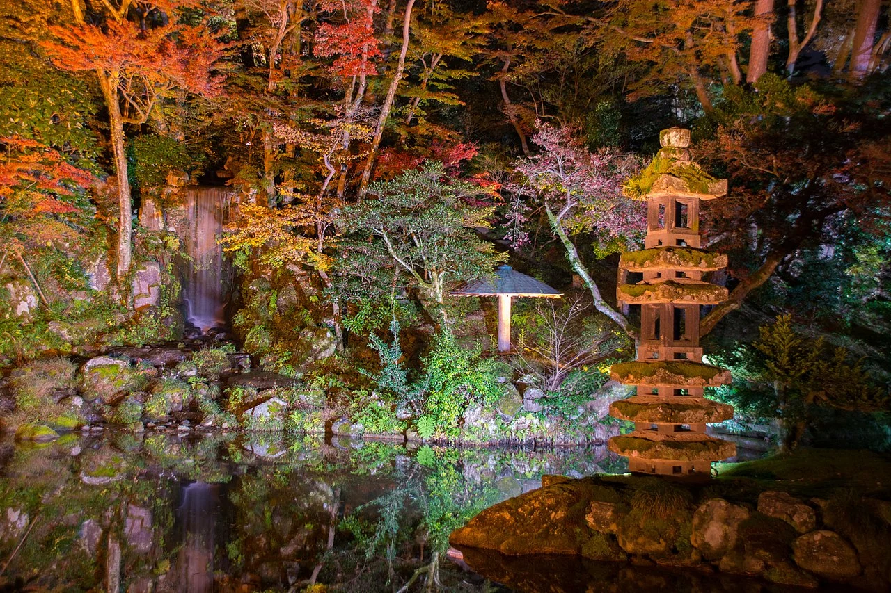 Red and yellow trees and a stone lantern reflected in water under nighttime lighting at Kenrokuen.